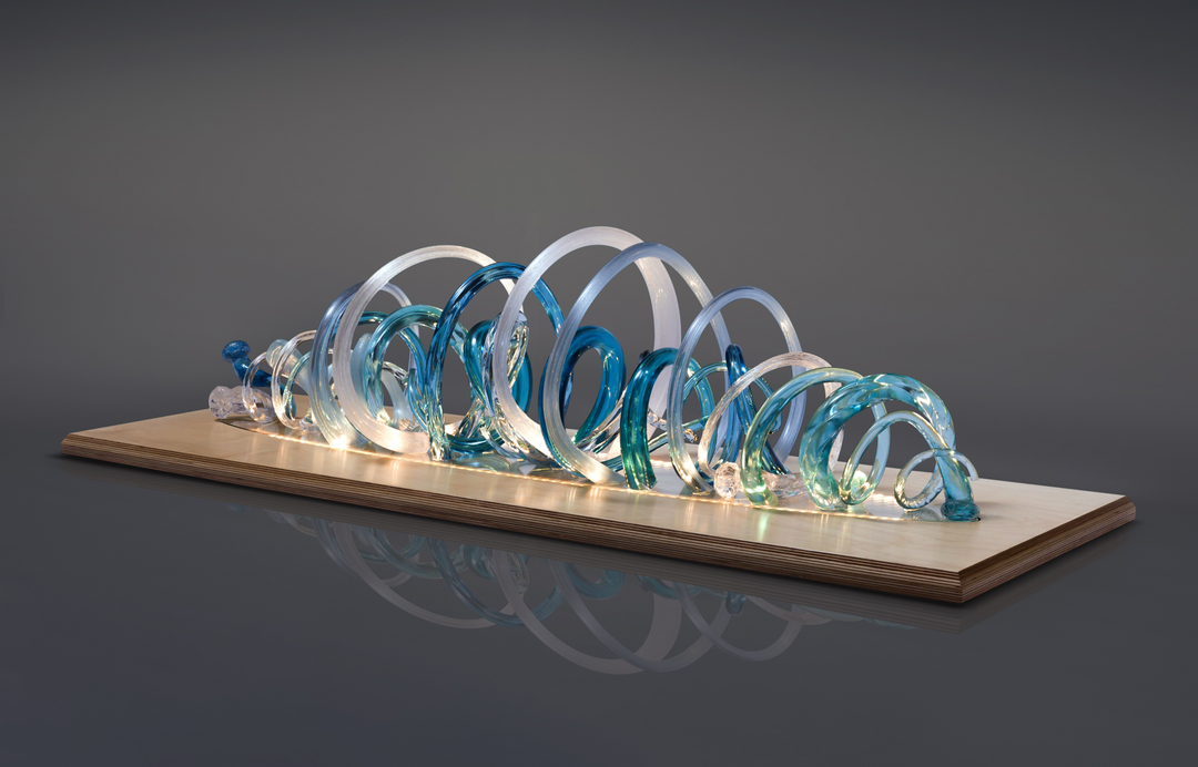 Glass sculpture of a wave, featuring flowing, ribbon-like forms in blue, aqua, and clear tones, set on a light wooden base.