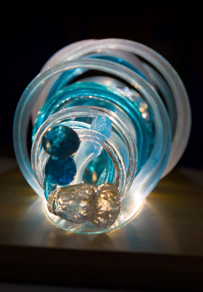 Glass sculpture of a wave, featuring flowing, ribbon-like forms in blue, aqua, and clear tones, set on a light wooden base.