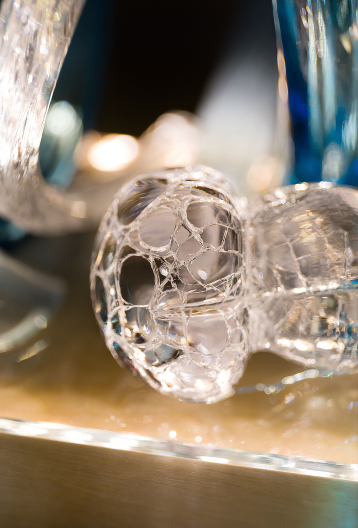 Glass sculpture of a wave, featuring flowing, ribbon-like forms in blue, aqua, and clear tones, set on a light wooden base.