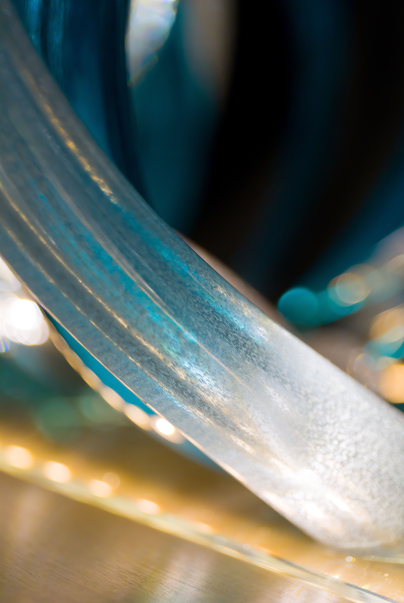 Glass sculpture of a wave, featuring flowing, ribbon-like forms in blue, aqua, and clear tones, set on a light wooden base.