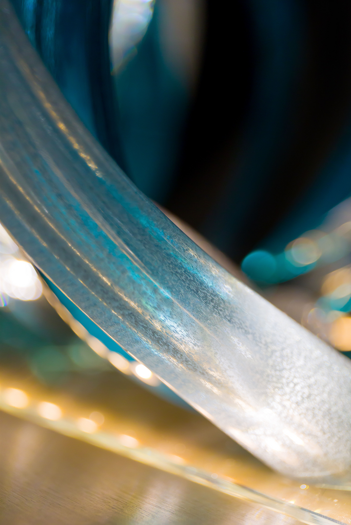 Glass sculpture of a wave, featuring flowing, ribbon-like forms in blue, aqua, and clear tones, set on a light wooden base.