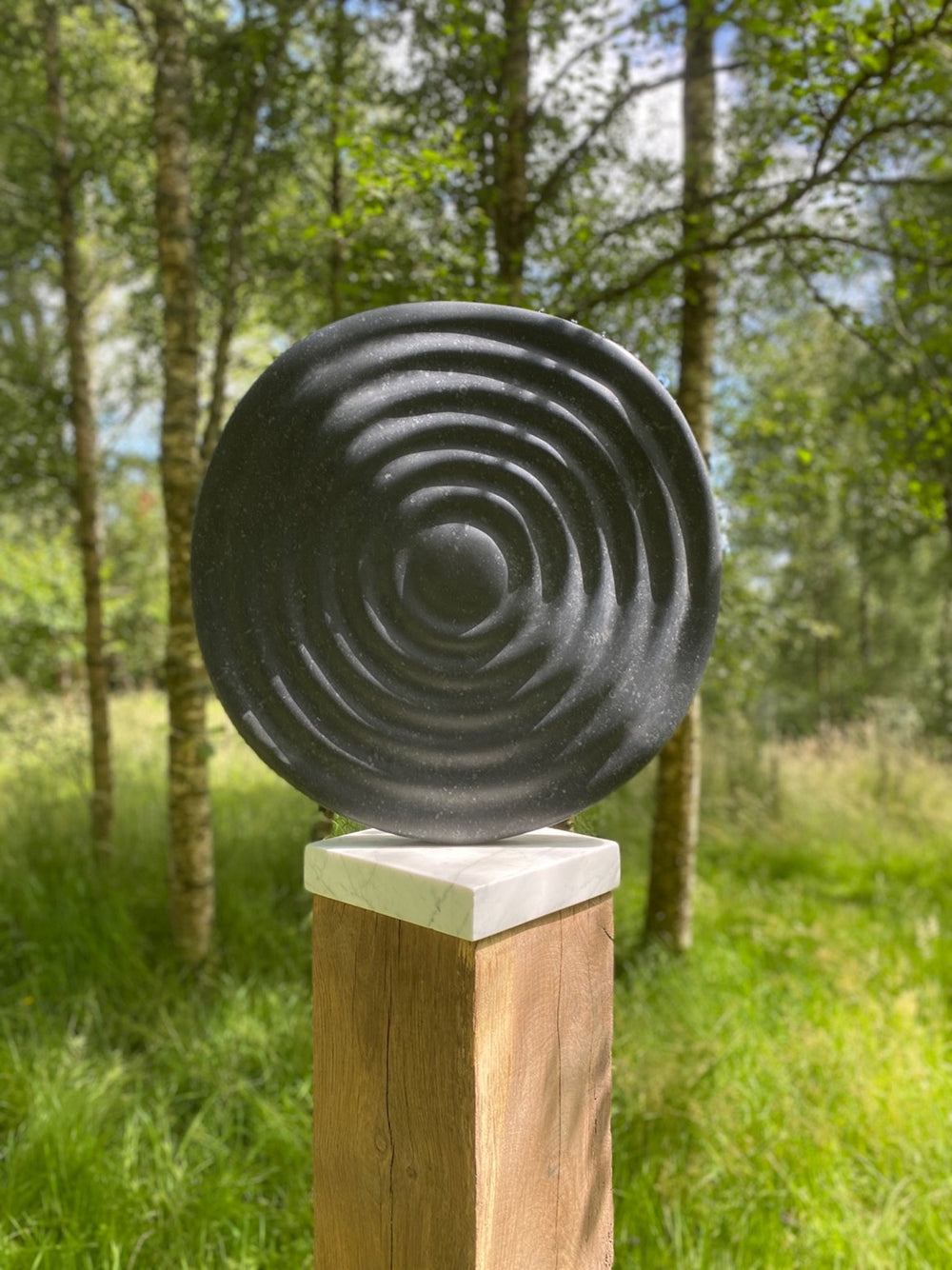 Circular abstract sculpture Wavelet – 2025 by Hamish Holman, carved in Kilkenny limestone with radiating ripple-like grooves across its dark stone surface, mounted on a white marble and oak plinth for indoor or outdoor display.