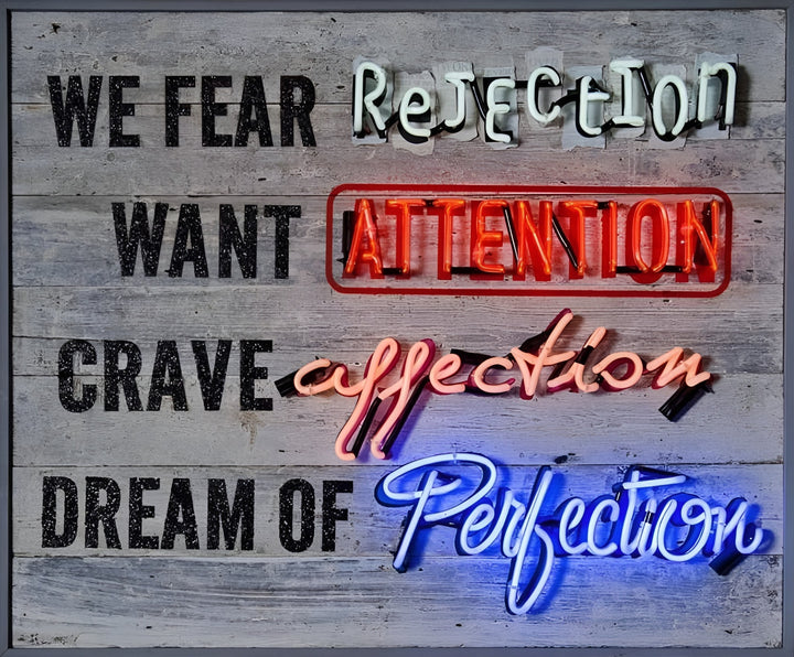 Neon artwork by Matthew Bracey with layered text reading: “WE FEAR Rejection, WANT ATTENTION, CRAVE affection, DREAM OF Perfection.” Each key word is highlighted in bold neon — “Rejection” in white, “ATTENTION” in red within a neon-outlined box, “affection” in pink, and “Perfection” in bright blue — all set against a weathered grey wooden background.