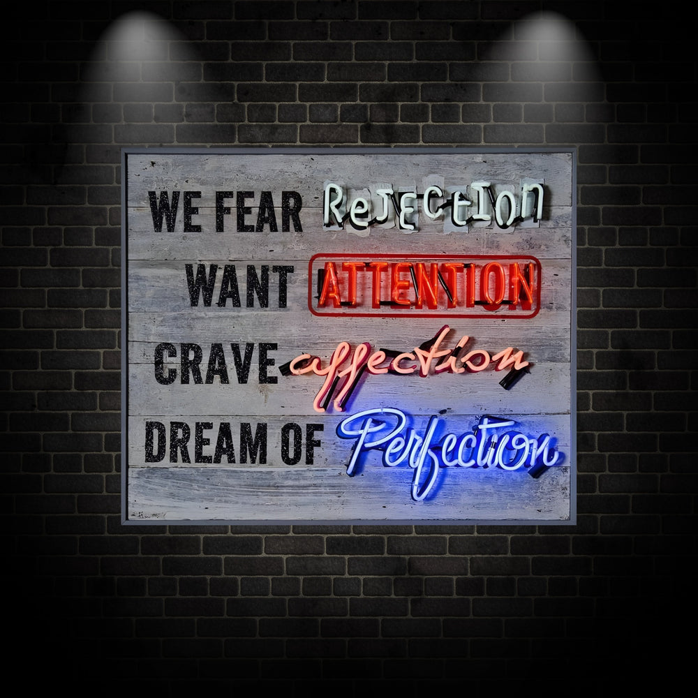 Neon artwork by Matthew Bracey with layered text reading: “WE FEAR Rejection, WANT ATTENTION, CRAVE affection, DREAM OF Perfection.” Each key word is highlighted in bold neon — “Rejection” in white, “ATTENTION” in red within a neon-outlined box, “affection” in pink, and “Perfection” in bright blue — all set against a weathered grey wooden background.