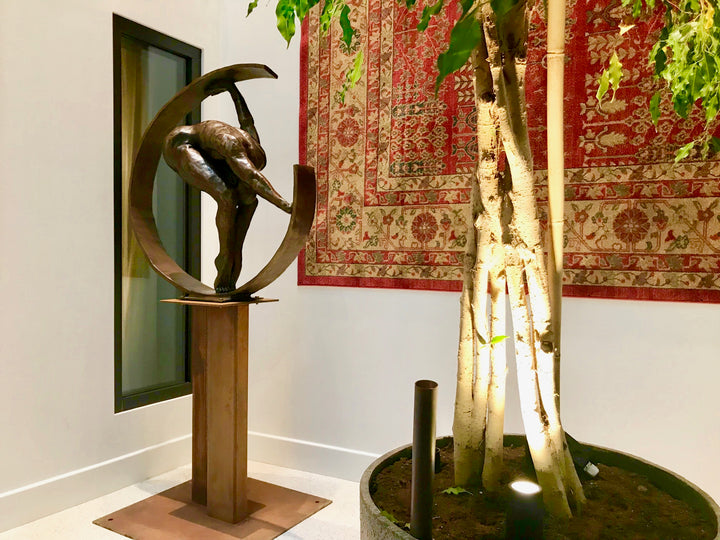 Weight of the World by Teresa Wells – a large, hand-cast bronze sculpture mounted on a tall steel base, depicting a nude male figure in a forward bend, gripping the ends of a sweeping curved bronze arc. The figure’s muscular back, bent legs, and tense feet convey physical strain and emotional weight, symbolising the burden of life’s pressures. Rich brown patina with subtle tonal variations enhances the anatomy and texture. Edition of 9, created 2019, 100cm × 45cm × 100cm, 100kg.
