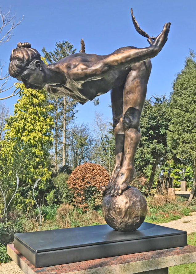 Ingenuity: Often thwarted by the shortsighted by Teresa Wells – a limited-edition bronze and steel sculpture depicting a poised, athletic female figure balanced on tiptoe atop a textured bronze sphere. The figure leans dramatically forward from the hips, arms extended behind and slightly upward, each hand delicately holding a detailed bronze feather. Muscular contours and tendons are rendered with lifelike precision, from the arched spine to the curled toes gripping the sphere. 