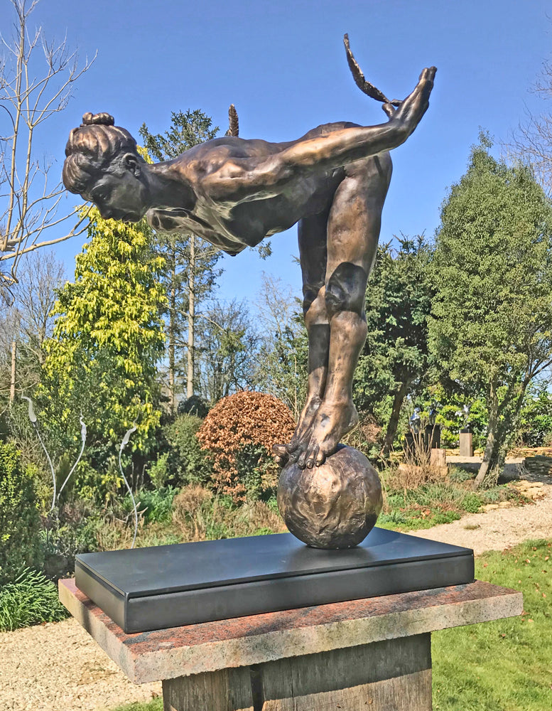 Ingenuity: Often thwarted by the shortsighted by Teresa Wells – a limited-edition bronze and steel sculpture depicting a poised, athletic female figure balanced on tiptoe atop a textured bronze sphere. The figure leans dramatically forward from the hips, arms extended behind and slightly upward, each hand delicately holding a detailed bronze feather. Muscular contours and tendons are rendered with lifelike precision, from the arched spine to the curled toes gripping the sphere. 