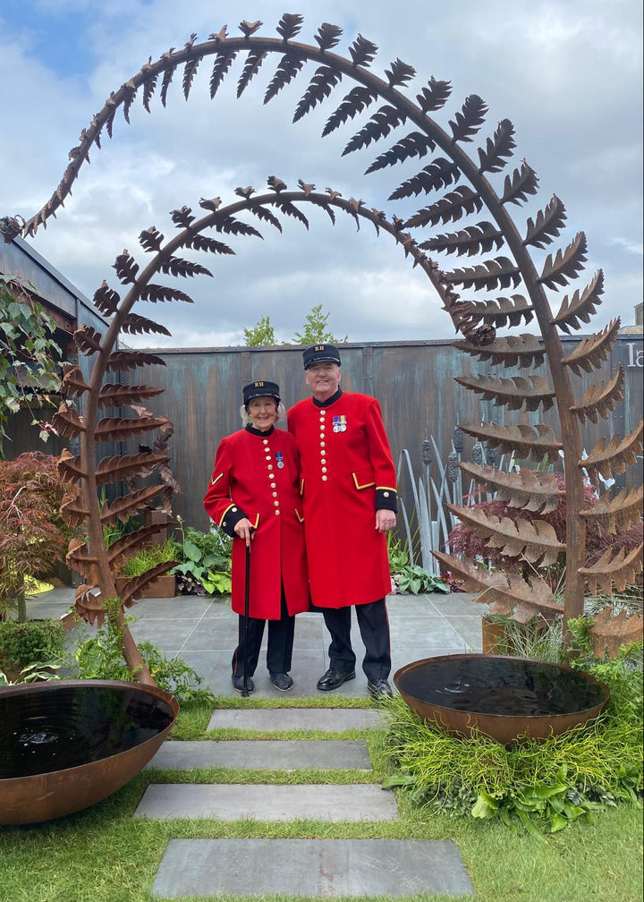 Towering corten steel fern sculptures form elegant archways within a lush garden landscape. Each sculptural frond, handcrafted from weathered steel, curves gracefully overhead and is designed as a water feature, releasing timed droplets into a pool below to create a tranquil, rhythmic ripple effect.