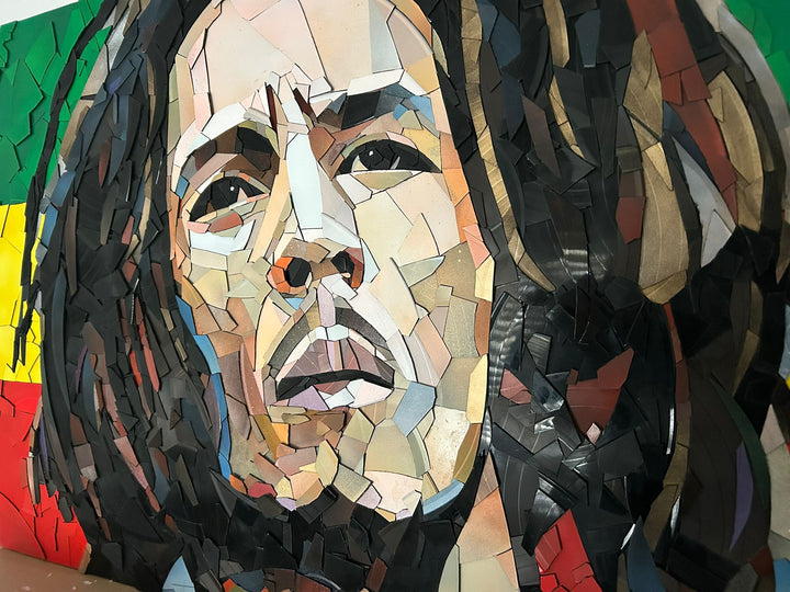 Ed Chapman Mosaic portrait of Bob Marley constructed from carefully cut fragments of reclaimed vinyl records, forming a highly detailed face with layered tonal variation and visible record grooves, set against a bold Rastafarian colour background that contrasts warm skin tones with strong graphic colour blocks and a textured, light-responsive surface.