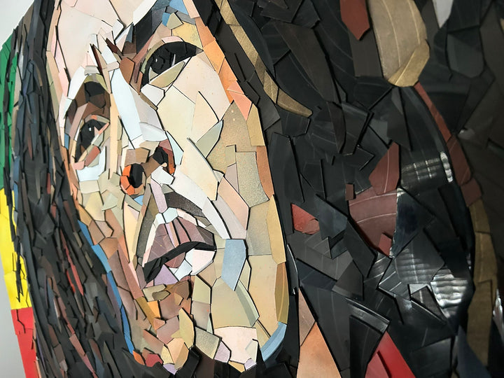 Ed Chapman Mosaic portrait of Bob Marley constructed from carefully cut fragments of reclaimed vinyl records, forming a highly detailed face with layered tonal variation and visible record grooves, set against a bold Rastafarian colour background that contrasts warm skin tones with strong graphic colour blocks and a textured, light-responsive surface.