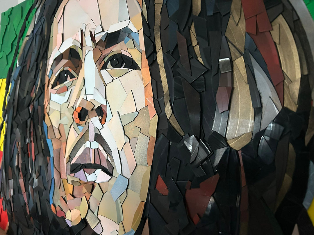 Ed Chapman Mosaic portrait of Bob Marley constructed from carefully cut fragments of reclaimed vinyl records, forming a highly detailed face with layered tonal variation and visible record grooves, set against a bold Rastafarian colour background that contrasts warm skin tones with strong graphic colour blocks and a textured, light-responsive surface.