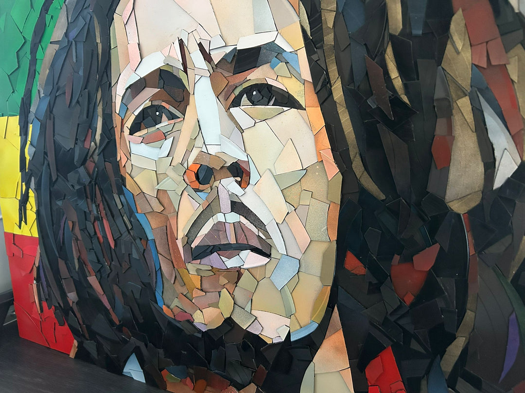 Ed Chapman Mosaic portrait of Bob Marley constructed from carefully cut fragments of reclaimed vinyl records, forming a highly detailed face with layered tonal variation and visible record grooves, set against a bold Rastafarian colour background that contrasts warm skin tones with strong graphic colour blocks and a textured, light-responsive surface.