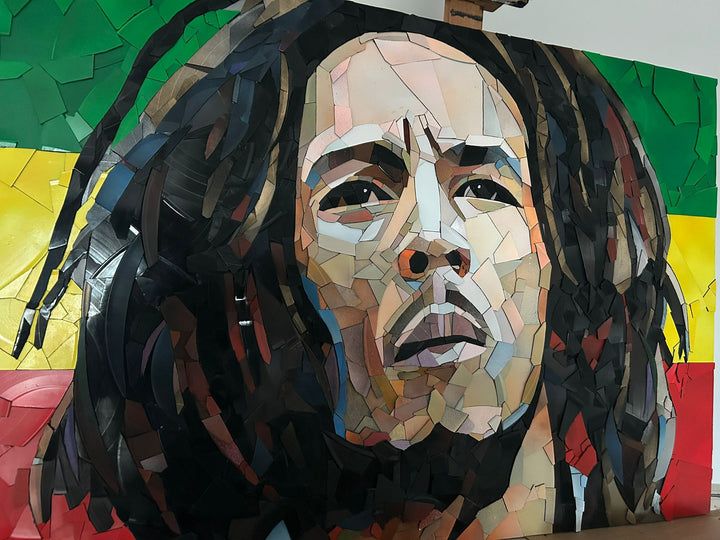 Ed Chapman Mosaic portrait of Bob Marley constructed from carefully cut fragments of reclaimed vinyl records, forming a highly detailed face with layered tonal variation and visible record grooves, set against a bold Rastafarian colour background that contrasts warm skin tones with strong graphic colour blocks and a textured, light-responsive surface.