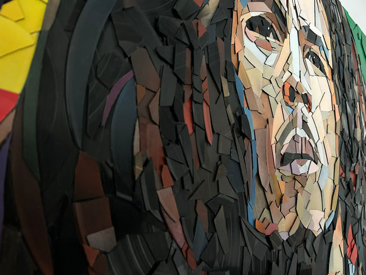 Ed Chapman Mosaic portrait of Bob Marley constructed from carefully cut fragments of reclaimed vinyl records, forming a highly detailed face with layered tonal variation and visible record grooves, set against a bold Rastafarian colour background that contrasts warm skin tones with strong graphic colour blocks and a textured, light-responsive surface.