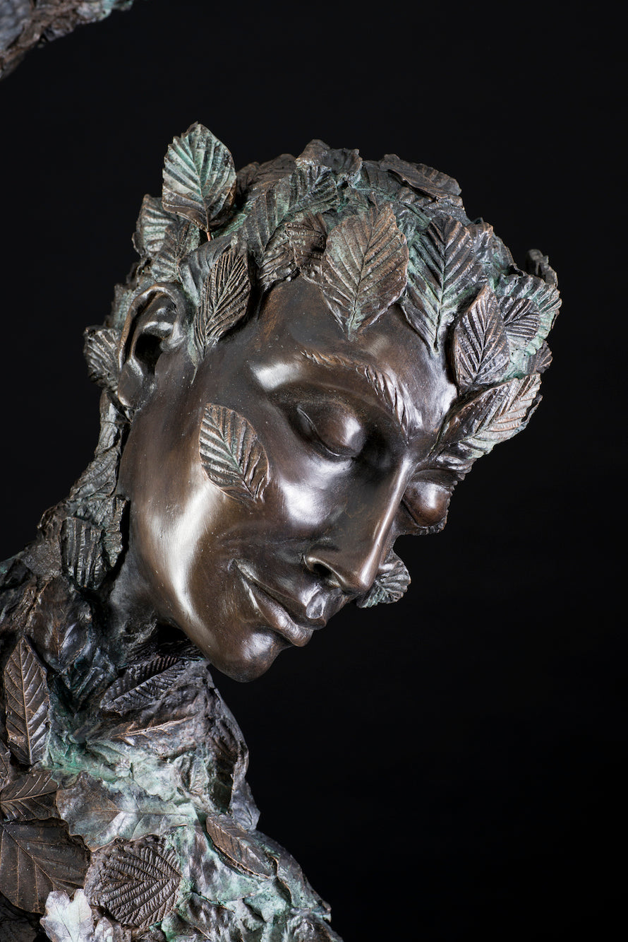 Large-scale patinated bronze sculpture of a stylised female figure emerging from a swirling, leaf-covered form. The composition appears wind-swept and dynamic, with detailed foliage enveloping the body and flowing into the air. Her serene face is partially obscured by leaves, symbolising transformation, movement, and harmony with nature. Rich in texture and emotional depth, this piece balances strength and sensitivity with a timeless organic elegance.