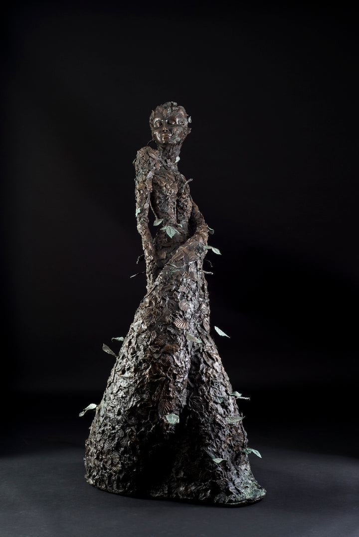 Tall patinated bronze sculpture of a female figure in a windswept, forward-leaning pose, emerging from a flowing base textured with detailed leaves. Her closed eyes and serene expression are partially obscured by sculpted foliage, symbolising a deep connection to nature and continual transformation. The figure’s dynamic form and elegant upward movement evoke renewal, seasonal change, and personal evolution. Rich bronze tones are enhanced by subtle green patination across the leafwork.