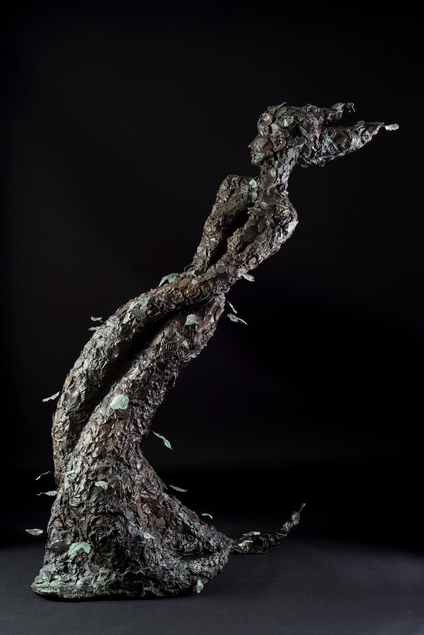 Tall patinated bronze sculpture of a female figure in a windswept, forward-leaning pose, emerging from a flowing base textured with detailed leaves. Her closed eyes and serene expression are partially obscured by sculpted foliage, symbolising a deep connection to nature and continual transformation. The figure’s dynamic form and elegant upward movement evoke renewal, seasonal change, and personal evolution. Rich bronze tones are enhanced by subtle green patination across the leafwork.