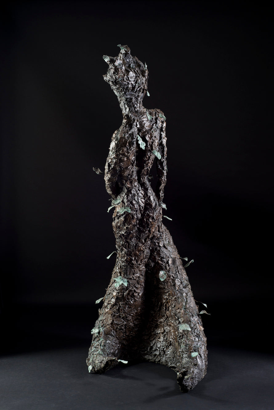 Tall patinated bronze sculpture of a female figure in a windswept, forward-leaning pose, emerging from a flowing base textured with detailed leaves. Her closed eyes and serene expression are partially obscured by sculpted foliage, symbolising a deep connection to nature and continual transformation. The figure’s dynamic form and elegant upward movement evoke renewal, seasonal change, and personal evolution. Rich bronze tones are enhanced by subtle green patination across the leafwork.