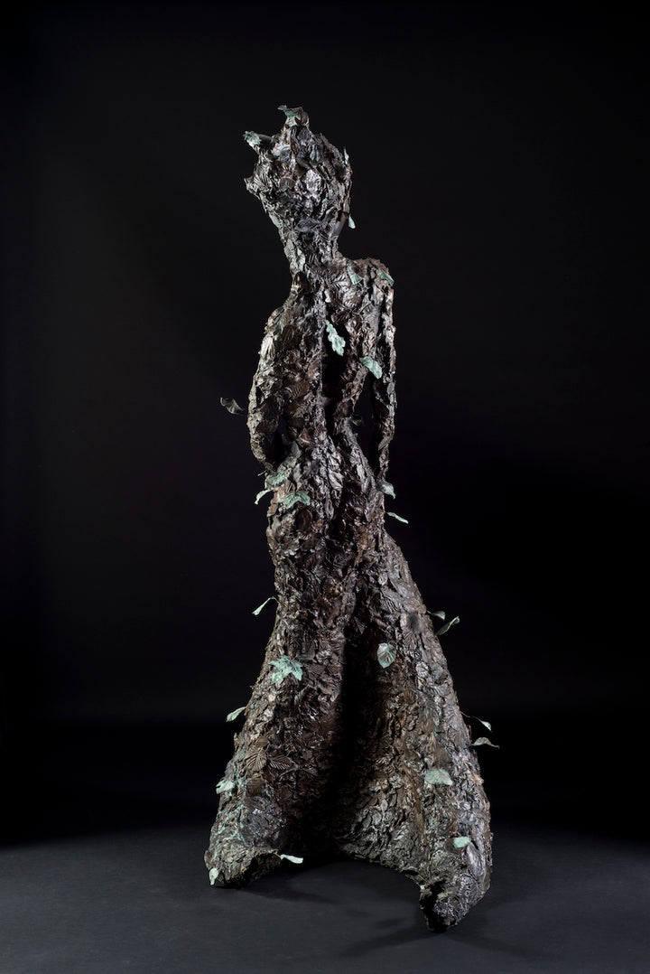 Tall patinated bronze sculpture of a female figure in a windswept, forward-leaning pose, emerging from a flowing base textured with detailed leaves. Her closed eyes and serene expression are partially obscured by sculpted foliage, symbolising a deep connection to nature and continual transformation. The figure’s dynamic form and elegant upward movement evoke renewal, seasonal change, and personal evolution. Rich bronze tones are enhanced by subtle green patination across the leafwork.