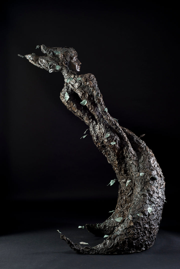 Tall patinated bronze sculpture of a female figure in a windswept, forward-leaning pose, emerging from a flowing base textured with detailed leaves. Her closed eyes and serene expression are partially obscured by sculpted foliage, symbolising a deep connection to nature and continual transformation. The figure’s dynamic form and elegant upward movement evoke renewal, seasonal change, and personal evolution. Rich bronze tones are enhanced by subtle green patination across the leafwork.