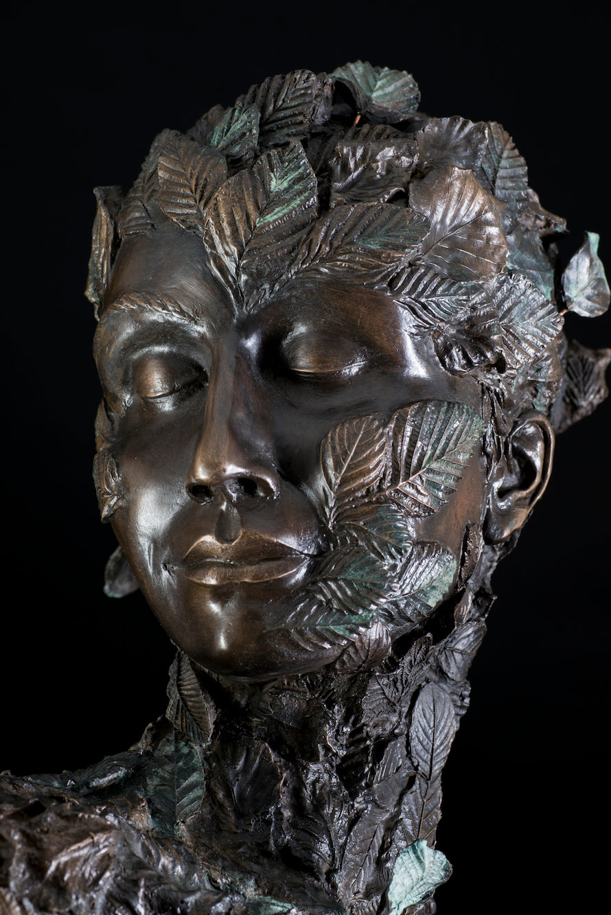 Tall patinated bronze sculpture of a female figure in a windswept, forward-leaning pose, emerging from a flowing base textured with detailed leaves. Her closed eyes and serene expression are partially obscured by sculpted foliage, symbolising a deep connection to nature and continual transformation. The figure’s dynamic form and elegant upward movement evoke renewal, seasonal change, and personal evolution. Rich bronze tones are enhanced by subtle green patination across the leafwork.
