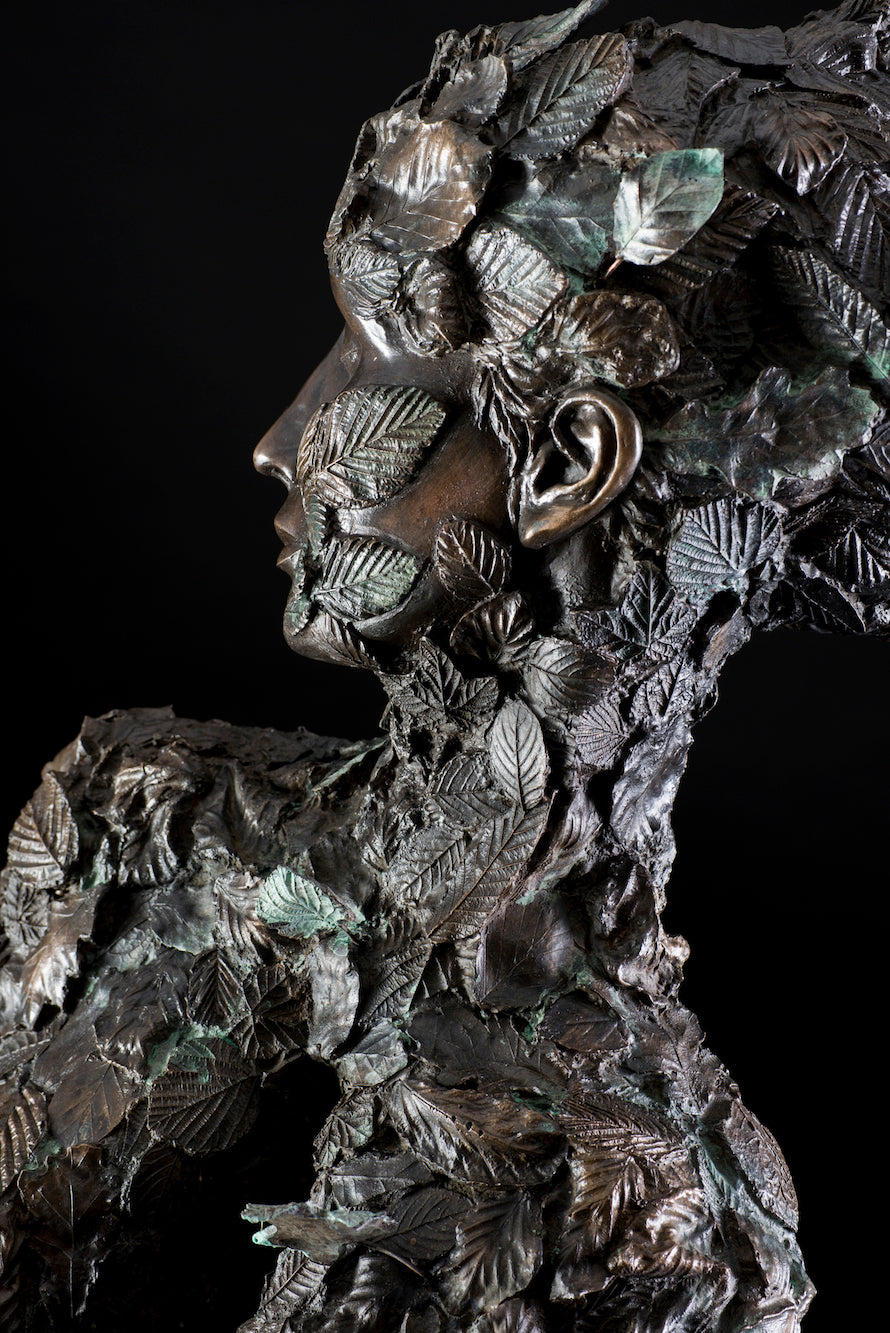 Tall patinated bronze sculpture of a female figure in a windswept, forward-leaning pose, emerging from a flowing base textured with detailed leaves. Her closed eyes and serene expression are partially obscured by sculpted foliage, symbolising a deep connection to nature and continual transformation. The figure’s dynamic form and elegant upward movement evoke renewal, seasonal change, and personal evolution. Rich bronze tones are enhanced by subtle green patination across the leafwork.