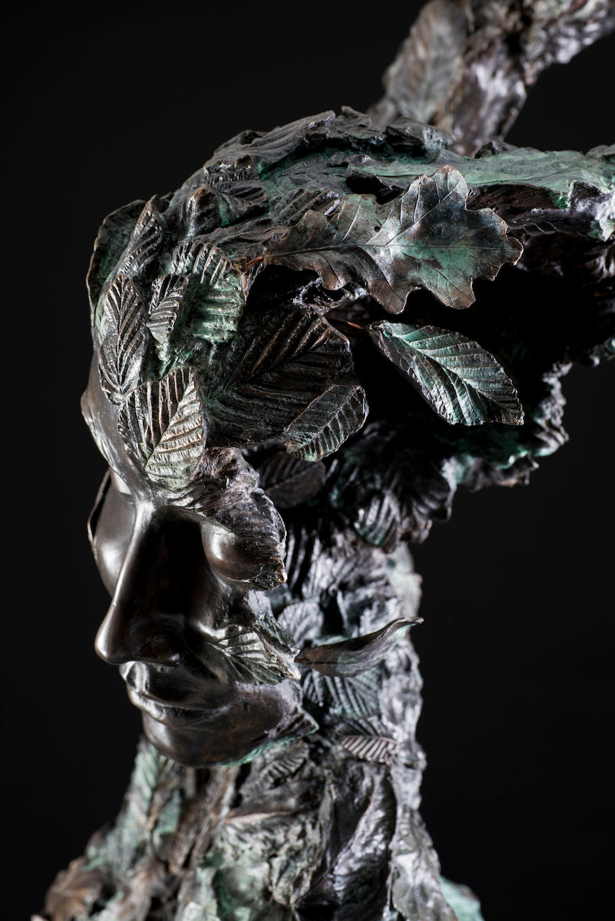 Tall patinated bronze sculpture of a female figure in a windswept, forward-leaning pose, emerging from a flowing base textured with detailed leaves. Her closed eyes and serene expression are partially obscured by sculpted foliage, symbolising a deep connection to nature and continual transformation. The figure’s dynamic form and elegant upward movement evoke renewal, seasonal change, and personal evolution. Rich bronze tones are enhanced by subtle green patination across the leafwork.