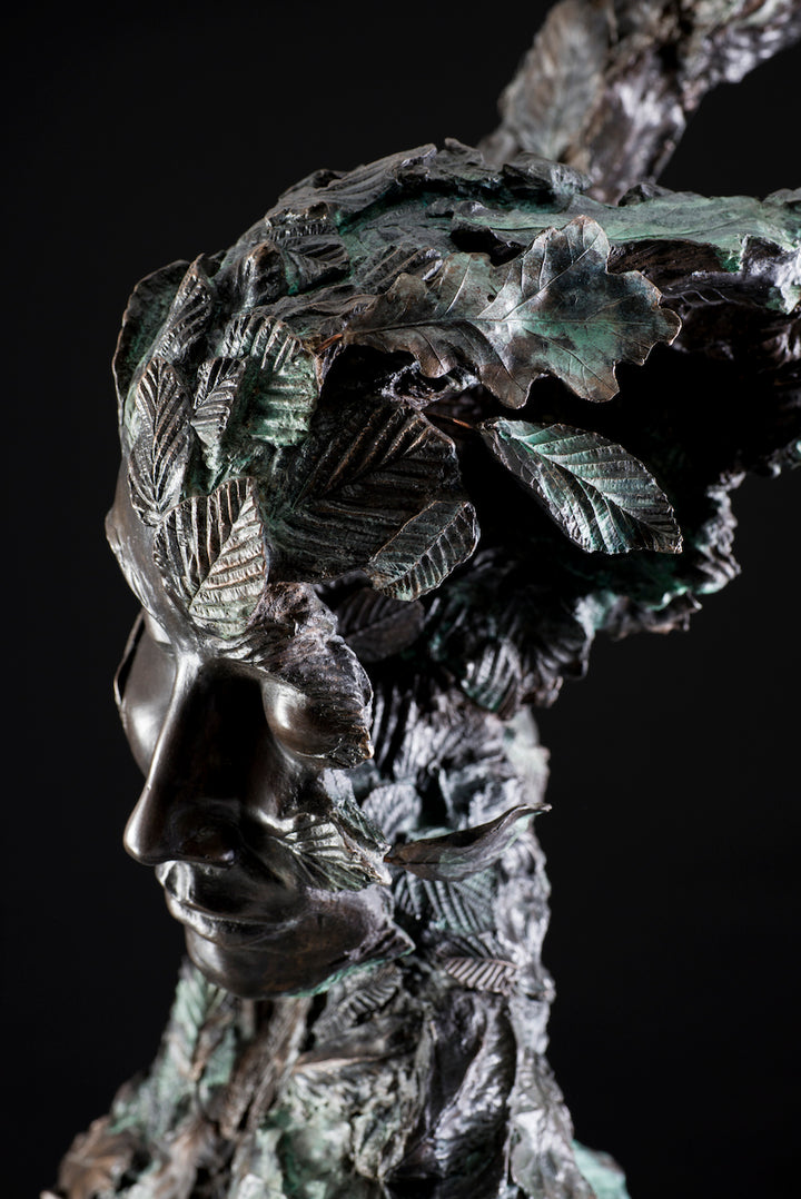 Tall patinated bronze sculpture of a female figure in a windswept, forward-leaning pose, emerging from a flowing base textured with detailed leaves. Her closed eyes and serene expression are partially obscured by sculpted foliage, symbolising a deep connection to nature and continual transformation. The figure’s dynamic form and elegant upward movement evoke renewal, seasonal change, and personal evolution. Rich bronze tones are enhanced by subtle green patination across the leafwork.