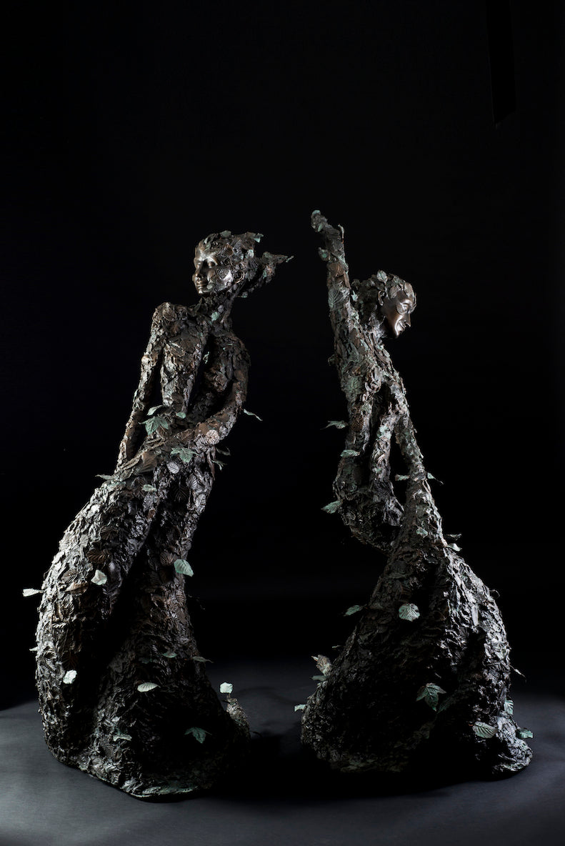 Tall patinated bronze sculpture of a female figure in a windswept, forward-leaning pose, emerging from a flowing base textured with detailed leaves. Her closed eyes and serene expression are partially obscured by sculpted foliage, symbolising a deep connection to nature and continual transformation. The figure’s dynamic form and elegant upward movement evoke renewal, seasonal change, and personal evolution. Rich bronze tones are enhanced by subtle green patination across the leafwork.