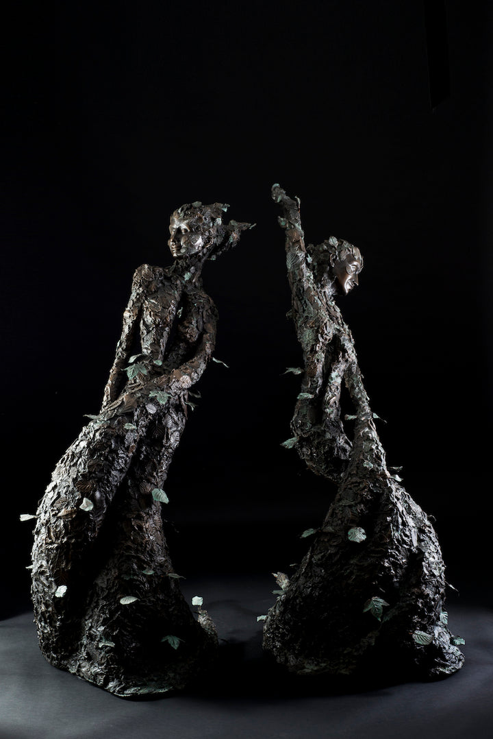 Tall patinated bronze sculpture of a female figure in a windswept, forward-leaning pose, emerging from a flowing base textured with detailed leaves. Her closed eyes and serene expression are partially obscured by sculpted foliage, symbolising a deep connection to nature and continual transformation. The figure’s dynamic form and elegant upward movement evoke renewal, seasonal change, and personal evolution. Rich bronze tones are enhanced by subtle green patination across the leafwork.