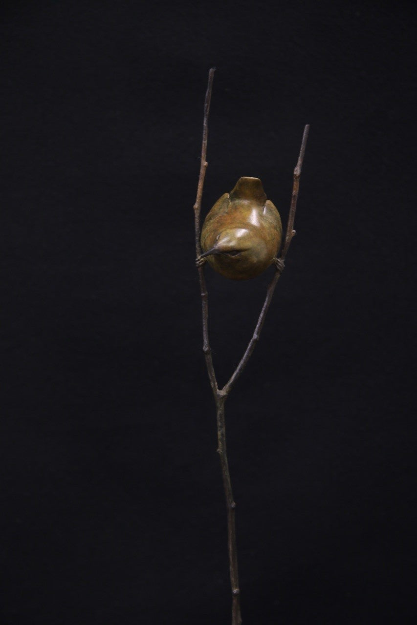 Bronze sculpture of a small bird perched between two delicate branches on a tall, slender stem, set against a black background.