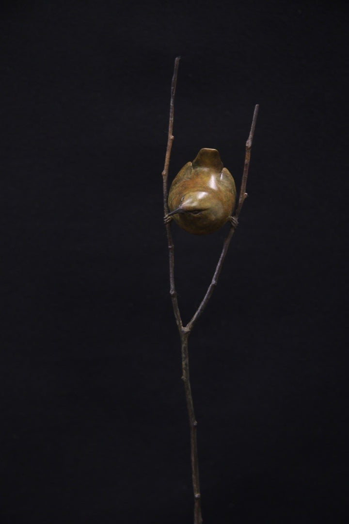 Bronze sculpture of a small bird perched between two delicate branches on a tall, slender stem, set against a black background.