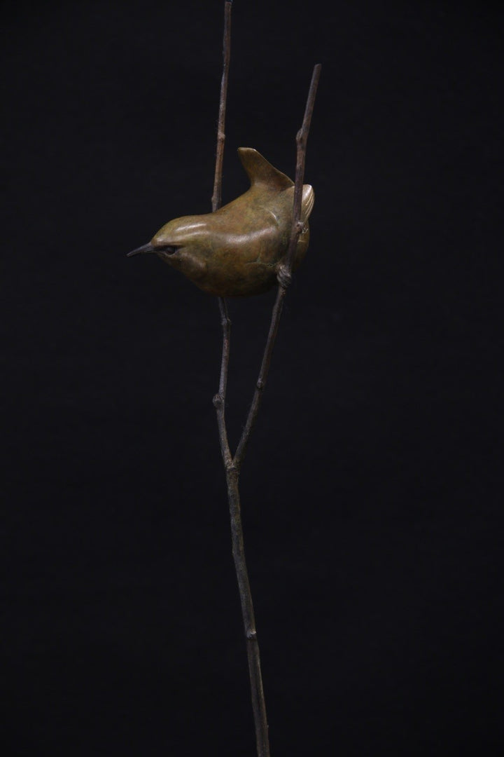 Bronze sculpture of a small bird perched between two delicate branches on a tall, slender stem, set against a black background.