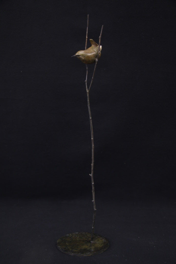 Bronze sculpture of a small bird perched between two delicate branches on a tall, slender stem, set against a black background.