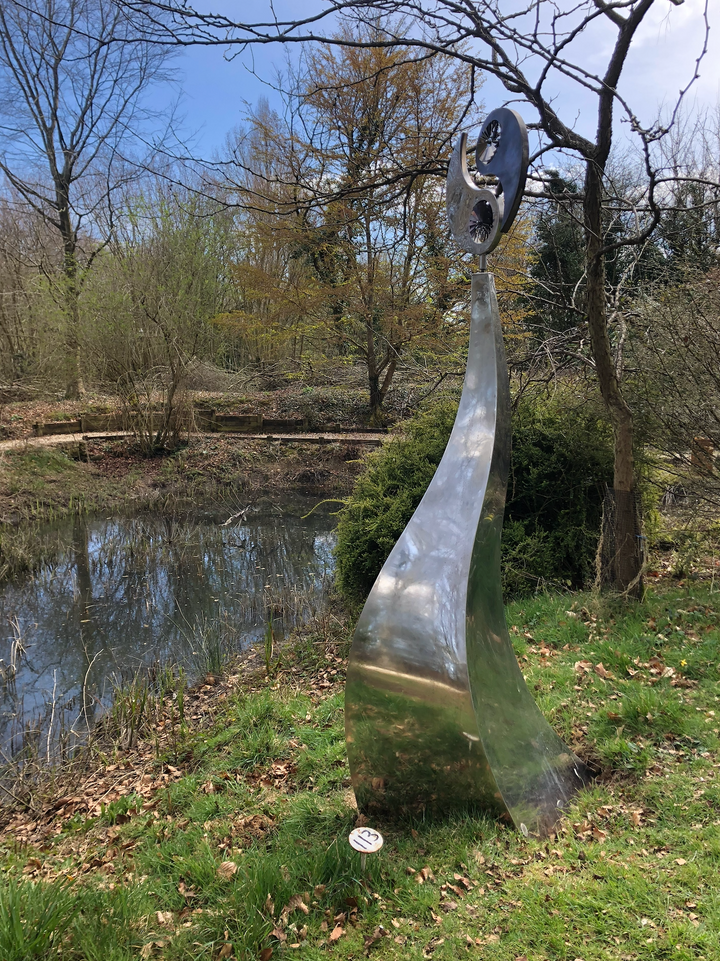 Polished stainless steel kinetic sculpture by Martin Jarvis featuring a tall, elegantly curved base topped with a dual-disc structure in motion. The upper element evokes a stylised yin-yang form, incorporating integrated fans that spin gently with the wind, creating a continuous, meditative rhythm.