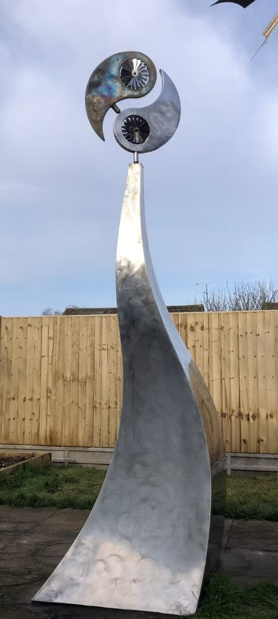 Polished stainless steel kinetic sculpture by Martin Jarvis featuring a tall, elegantly curved base topped with a dual-disc structure in motion. The upper element evokes a stylised yin-yang form, incorporating integrated fans that spin gently with the wind, creating a continuous, meditative rhythm.