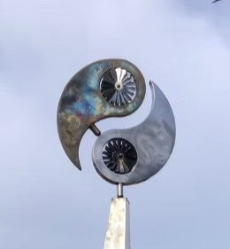 Polished stainless steel kinetic sculpture by Martin Jarvis featuring a tall, elegantly curved base topped with a dual-disc structure in motion. The upper element evokes a stylised yin-yang form, incorporating integrated fans that spin gently with the wind, creating a continuous, meditative rhythm.

