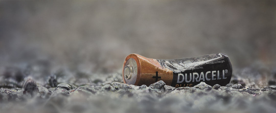 Simon Taylor, YOU LEFT ME POWERLESS (Original), oil and acrylic on aluminium, 25 x 60cm – photorealist painting of a discarded Duracell battery lying on grey gravel, rendered with hyperreal precision.