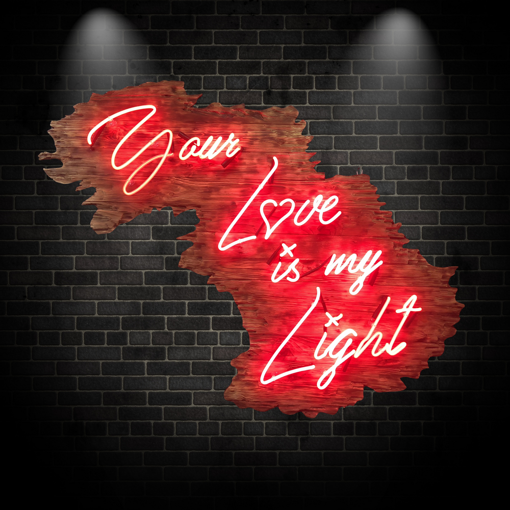 Neon artwork by Matthew Bracey in warm white script reading “Your Love is my Light,” mounted on a uniquely contoured, deep red wood panel shaped like a stylised landscape or abstract map. The word “Love” is stylised with a heart-shaped “o,” glowing against rich wood grain and glowing red backlighting.