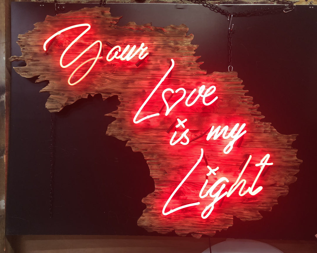 Neon artwork by Matthew Bracey in warm white script reading “Your Love is my Light,” mounted on a uniquely contoured, deep red wood panel shaped like a stylised landscape or abstract map. The word “Love” is stylised with a heart-shaped “o,” glowing against rich wood grain and glowing red backlighting.