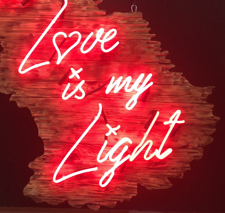 Neon artwork by Matthew Bracey in warm white script reading “Your Love is my Light,” mounted on a uniquely contoured, deep red wood panel shaped like a stylised landscape or abstract map. The word “Love” is stylised with a heart-shaped “o,” glowing against rich wood grain and glowing red backlighting.