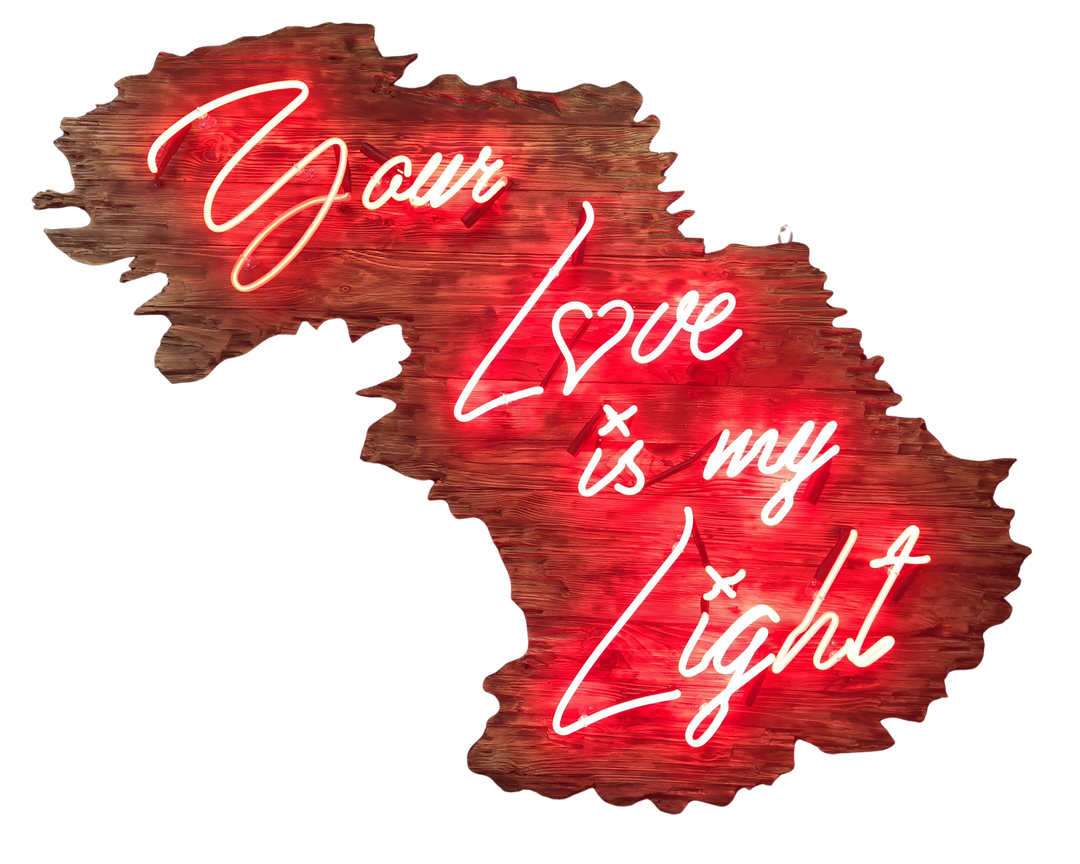 Neon artwork by Matthew Bracey in warm white script reading “Your Love is my Light,” mounted on a uniquely contoured, deep red wood panel shaped like a stylised landscape or abstract map. The word “Love” is stylised with a heart-shaped “o,” glowing against rich wood grain and glowing red backlighting.