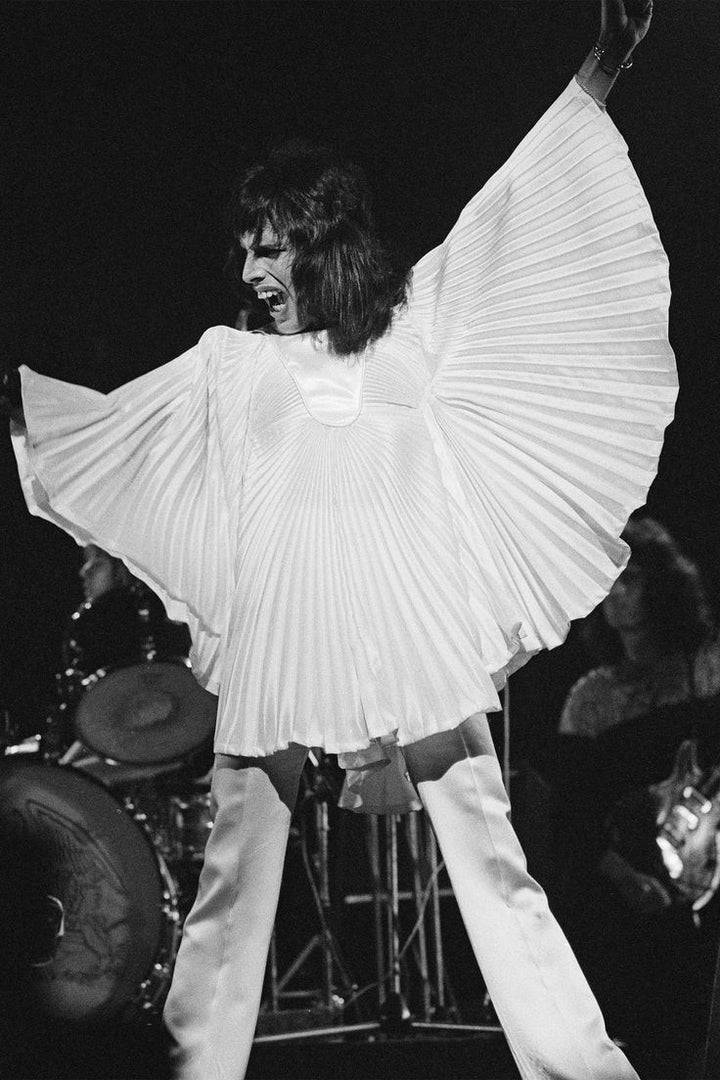 Freddie Mercury performing on stage in a dramatic white pleated top designed by Dame Zandra Rhodes, captured mid-song with a microphone in hand and theatrical lighting highlighting the garment’s sculptural folds.