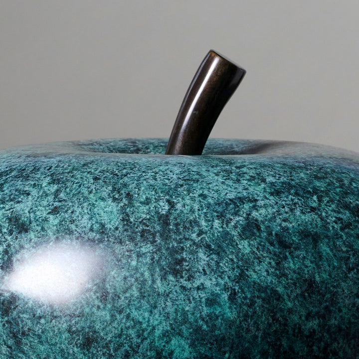 Blue-green patinated bronze apple sculpture by James Parker, positioned on a natural stone boulder within a landscaped garden setting.
