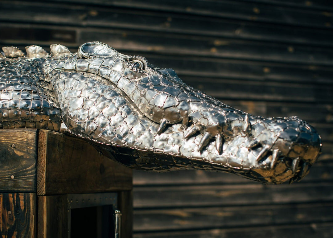 ‘Crocodile on a Box’ by Michael Turner, a unique, one-of-one stainless steel wildlife sculpture depicting a life-size crocodile resting across a large wooden crate. Handcrafted from intricately shaped and welded stainless steel, the crocodile features individually detailed silver scales, powerful jaws, raised ridges along its back, muscular legs and a long tail extending over the edge. The rustic brown timber box features a metal hatch, locking mechanism and bold black DANGER lettering, creating a striking 