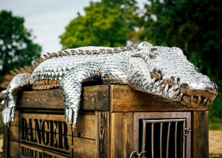 ‘Crocodile on a Box’ by Michael Turner, a unique, one-of-one stainless steel wildlife sculpture depicting a life-size crocodile resting across a large wooden crate. Handcrafted from intricately shaped and welded stainless steel, the crocodile features individually detailed silver scales, powerful jaws, raised ridges along its back, muscular legs and a long tail extending over the edge. The rustic brown timber box features a metal hatch, locking mechanism and bold black DANGER lettering, creating a striking 