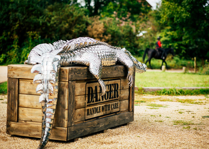 ‘Crocodile on a Box’ by Michael Turner, a unique, one-of-one stainless steel wildlife sculpture depicting a life-size crocodile resting across a large wooden crate. Handcrafted from intricately shaped and welded stainless steel, the crocodile features individually detailed silver scales, powerful jaws, raised ridges along its back, muscular legs and a long tail extending over the edge. The rustic brown timber box features a metal hatch, locking mechanism and bold black DANGER lettering, creating a striking 