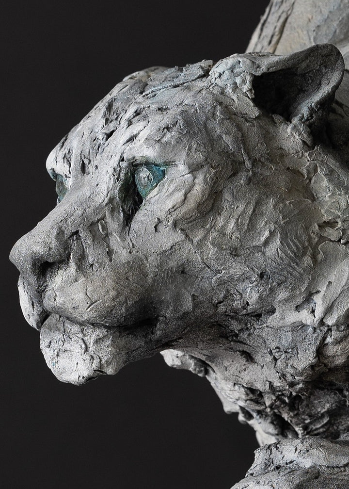 Sculpture of a stalking snow leopard by Lucy d’Auvergne, created in cast slate with raw earth pigments, showing a powerful crouched pose with textured grey and earthy tones, green eyes, and mounted on a granite base.