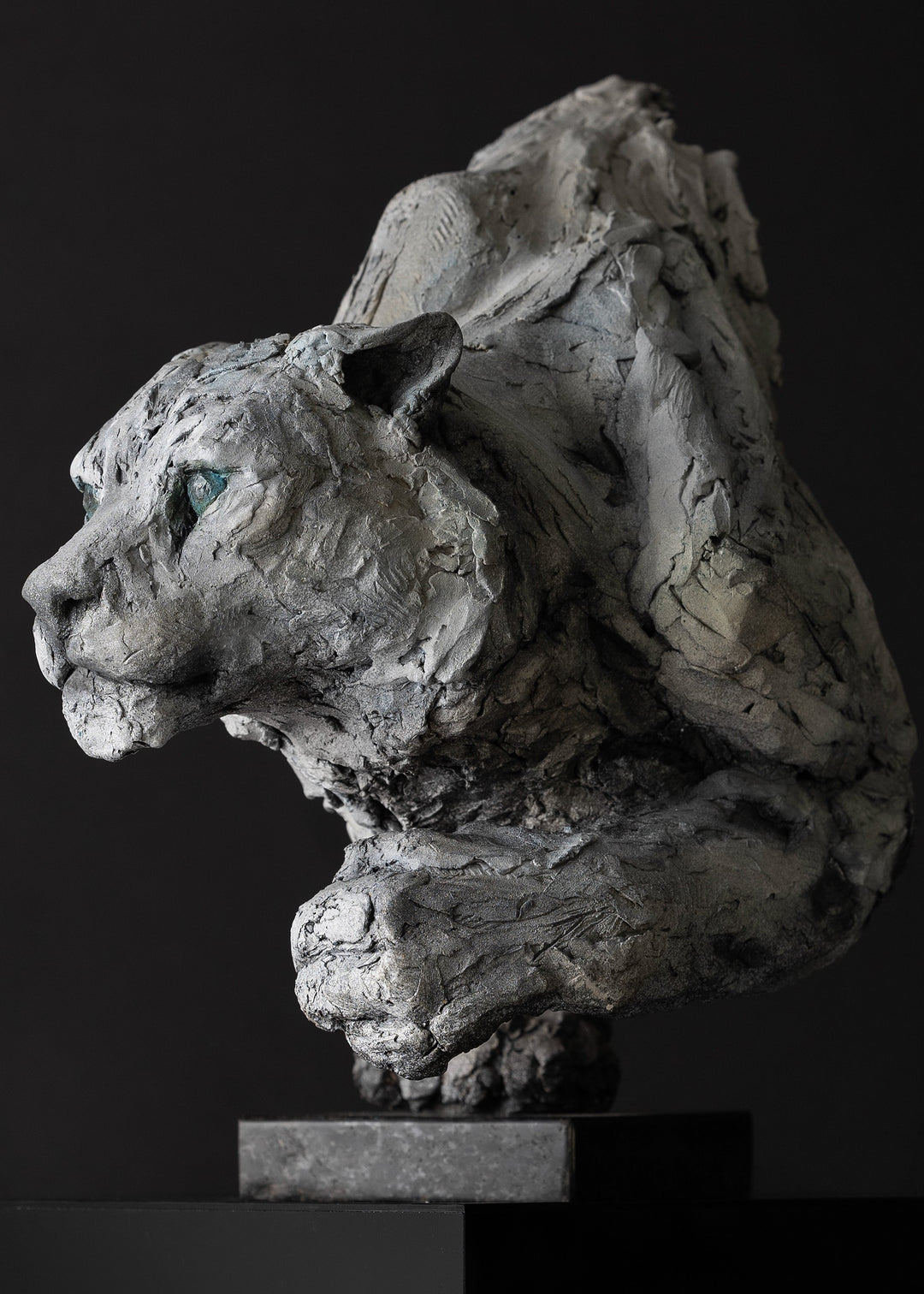 Sculpture of a stalking snow leopard by Lucy d’Auvergne, created in cast slate with raw earth pigments, showing a powerful crouched pose with textured grey and earthy tones, green eyes, and mounted on a granite base.