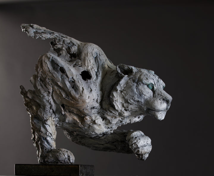 Sculpture of a stalking snow leopard by Lucy d’Auvergne, created in cast slate with raw earth pigments, showing a powerful crouched pose with textured grey and earthy tones, green eyes, and mounted on a granite base.